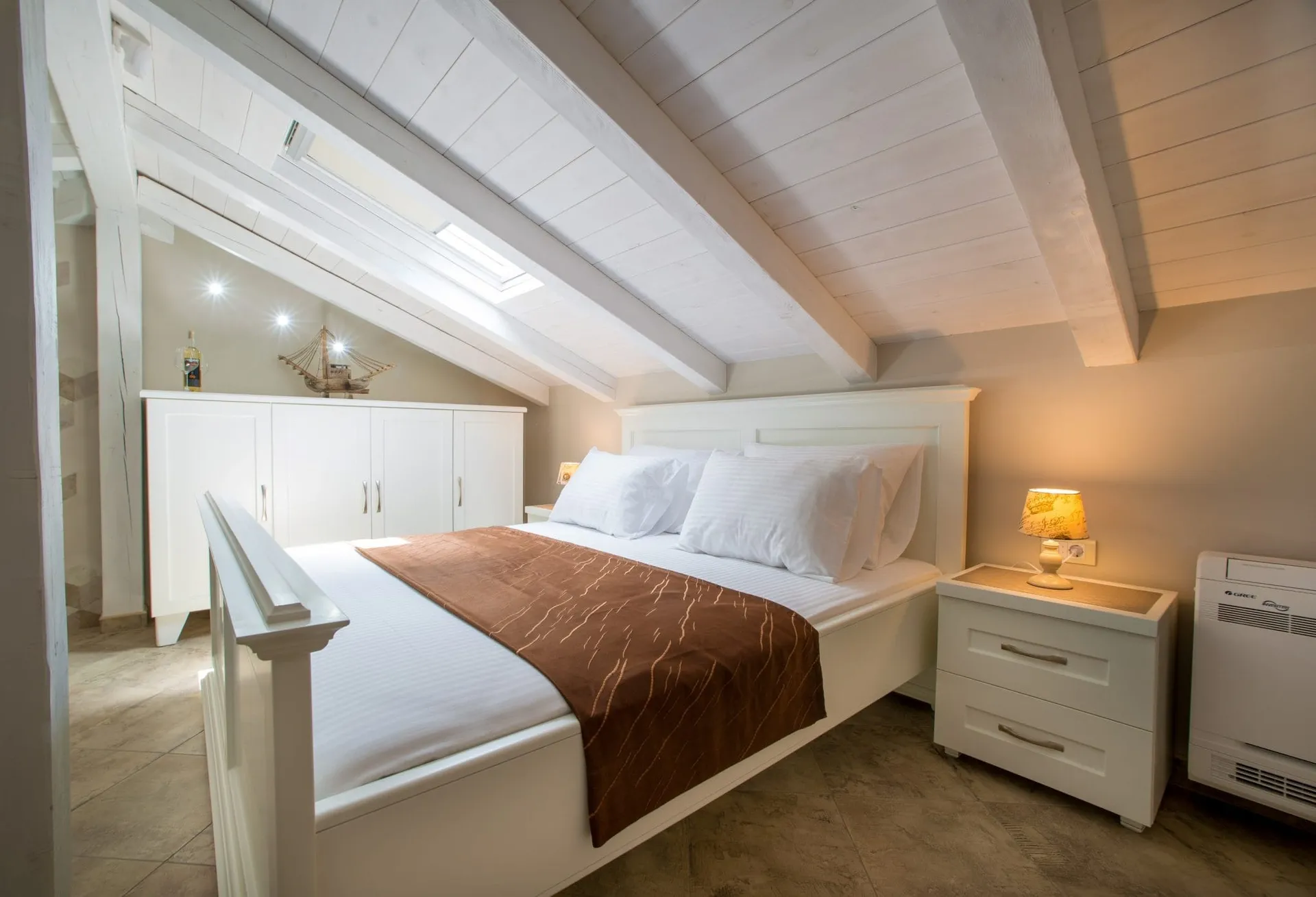 Dachschlafzimmer mit weißem Bett, Holzbalken, Dachfenster und weißen Möbeln.
