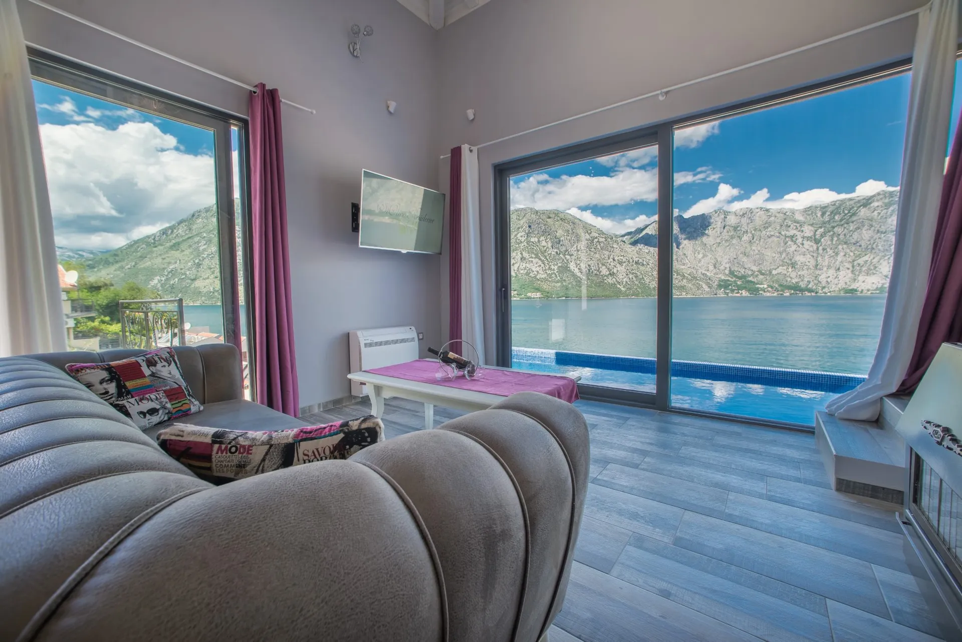 Wohnzimmer mit großen Fenstern mit Blick auf einen See und Berge, einem Sofa und einem wandmontierten Fernseher.