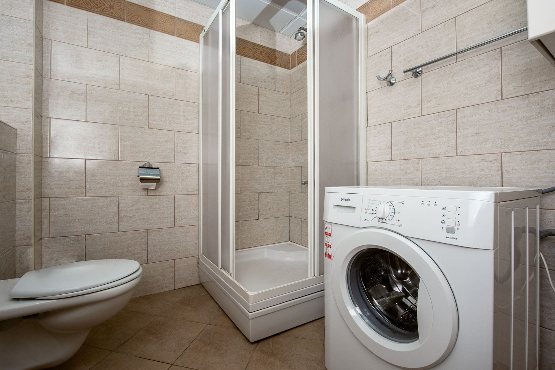 Badezimmer in einer Wohnung in Bečići, Montenegro: Duschkabine, Toilette und Gorenje-Waschmaschine, beige Fliesen.
