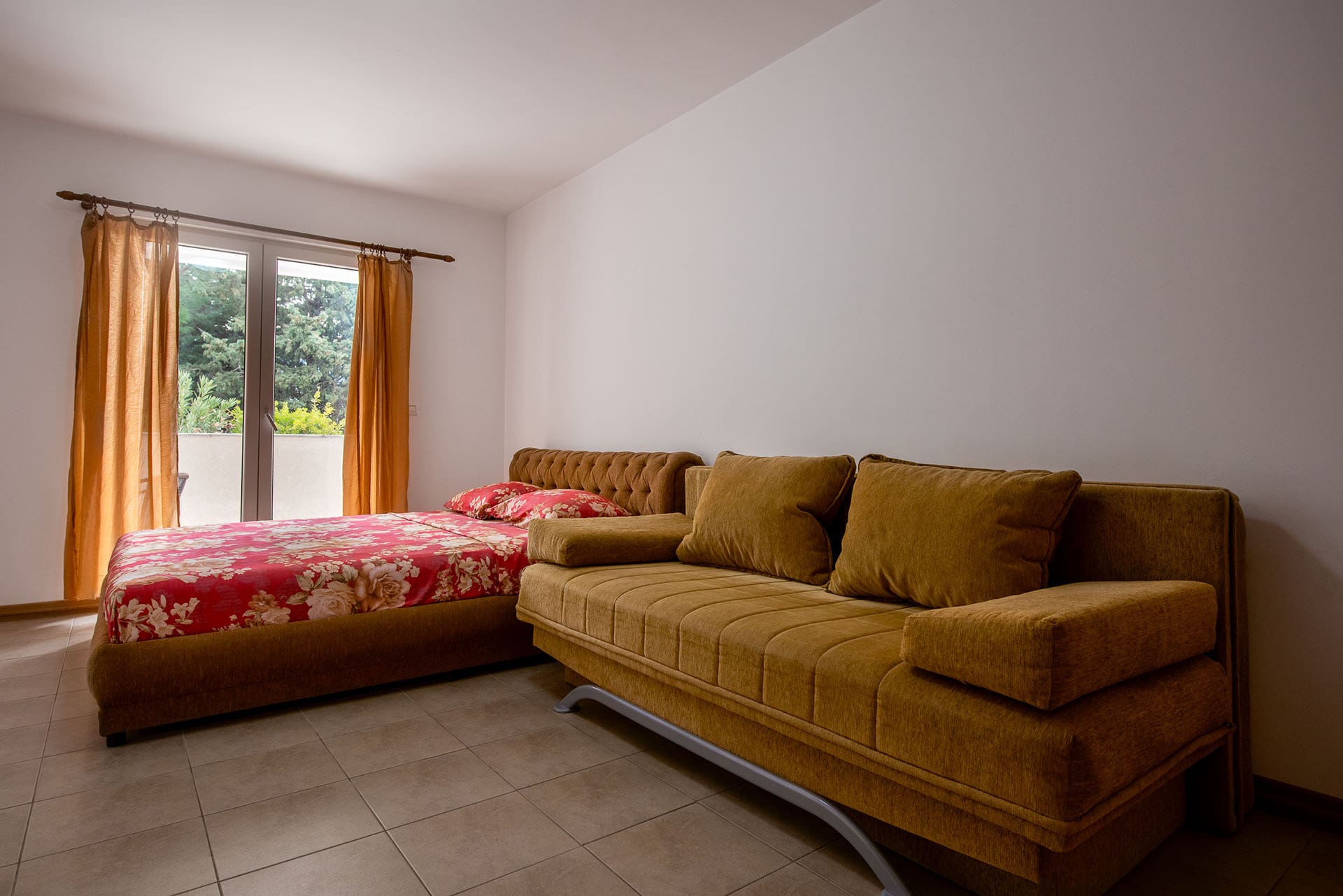 Schlafzimmer in einer Wohnung in Bečići, Montenegro: Doppelbett, senfgelbes Sofa und Zugang zum Balkon mit Blick ins Grüne.