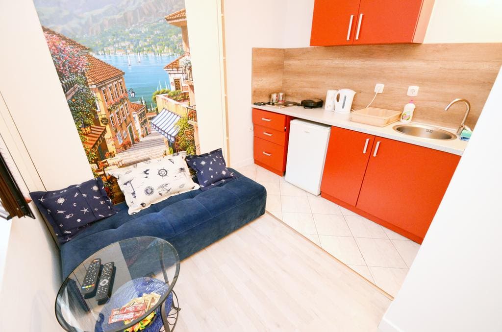 Wohnzimmer mit Küche in einem Haus in Kotor, Montenegro: blaues Sofa, helle Küchenzeile und Fototapete mit Meereslandschaft.