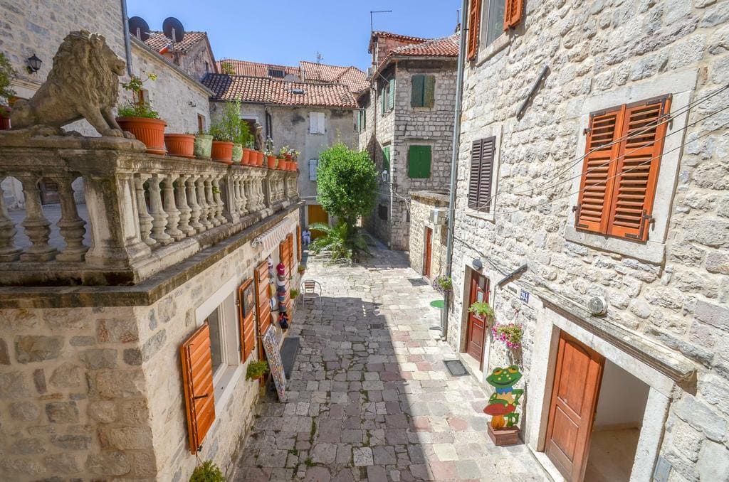 Enge gepflasterte Gasse in der Altstadt von Kotor, Montenegro, mit Steinhäusern, Fensterläden und einer Terrasse mit Löwenstatue.