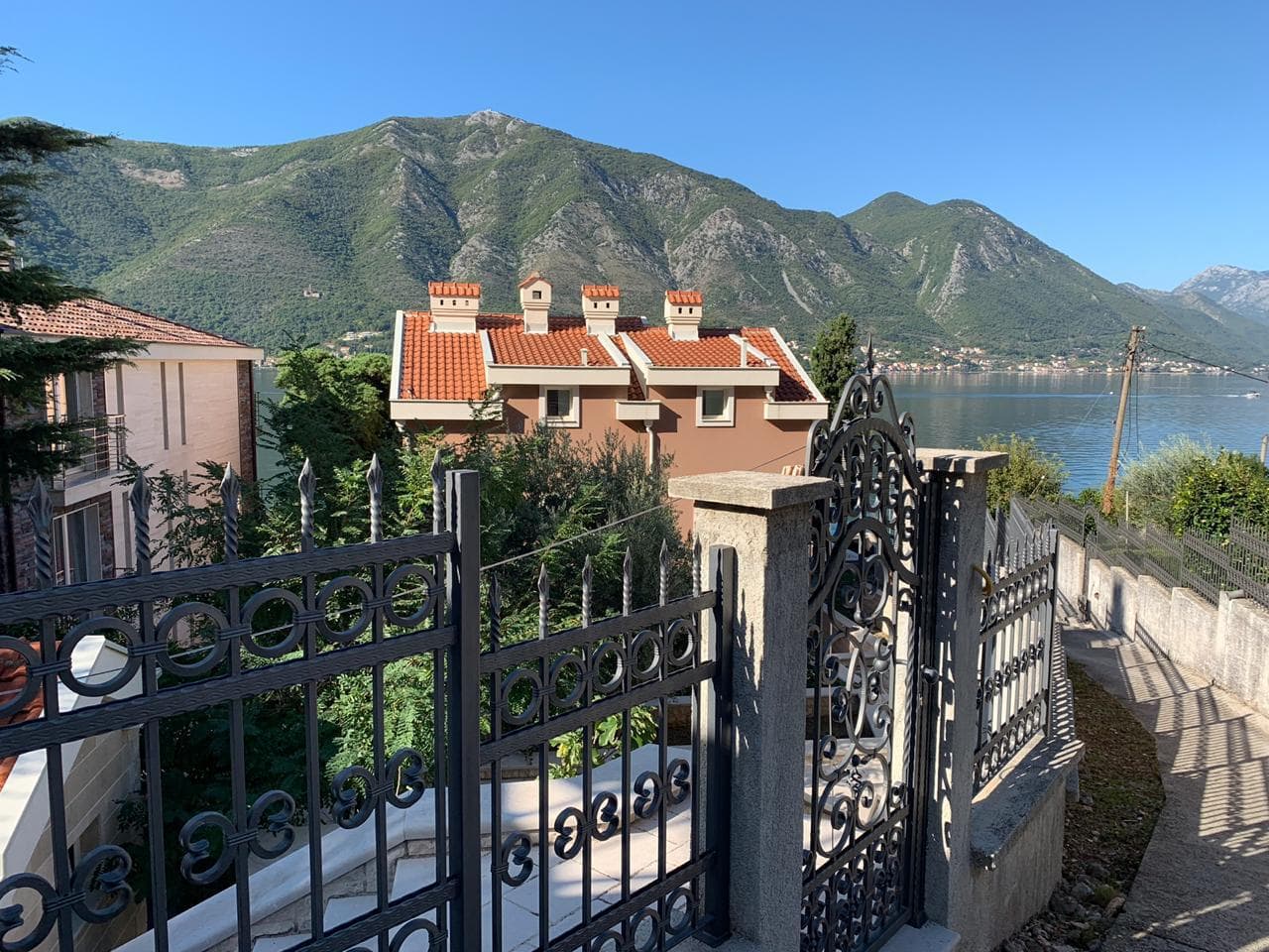 Pogled na vilu u Dobroti, Crna Gora: kovana ograda, terakotni krov i panoramski pogled na Bokokotorski zaliv i planine.