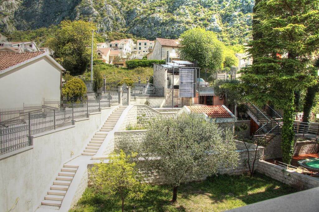 Dvorište vile u Dobroti, Crna Gora: kamene stepenice, zelena bašta sa maslinom i pogled na planine.
