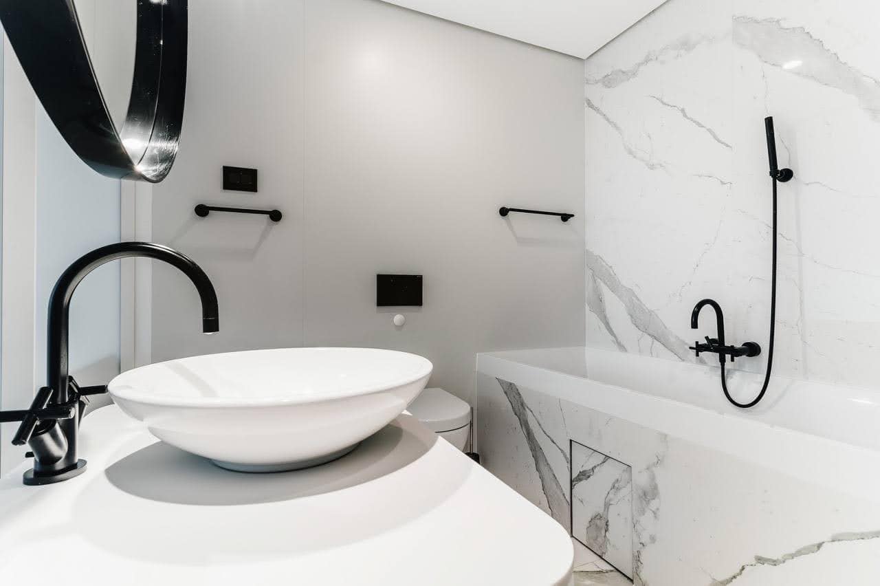 Karadağ, Budva’daki bir dairede modern banyo: mermer görünümlü kaplama, siyah armatürler ve tezgah üstü lavabo.