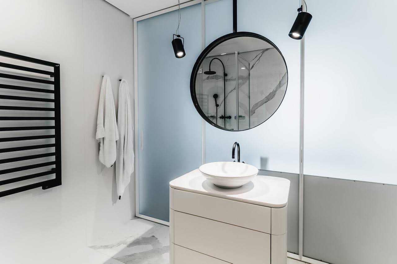Karadağ, Budva’daki bir dairede modern banyo: yuvarlak ayna, tezgah üstü lavabo ve mat bölmeler.