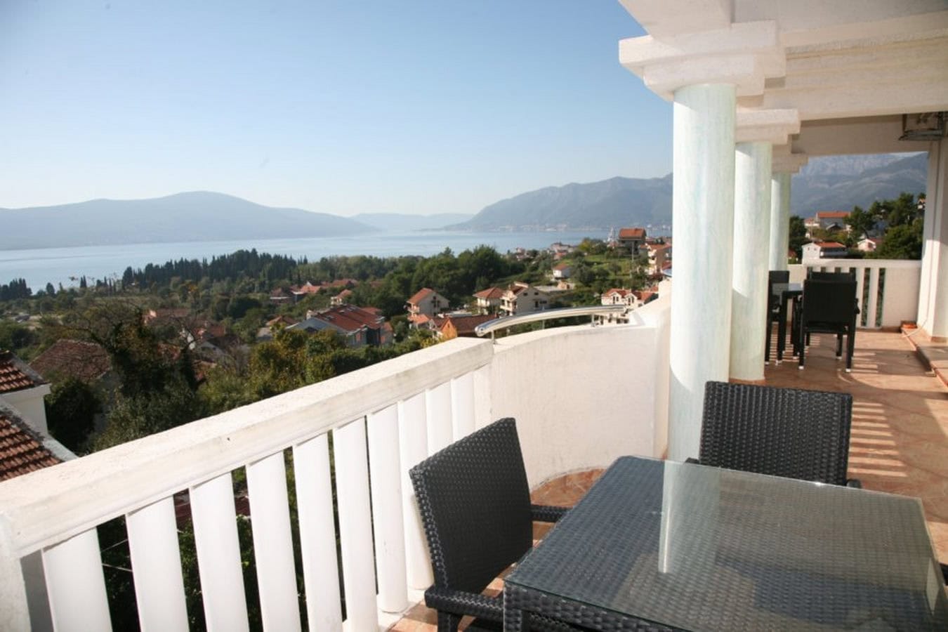 Terrasse der Villa in Tivat, Montenegro: Meer- und Bergblick, geflochtene Möbel mit Glastisch, weiße Säulen.