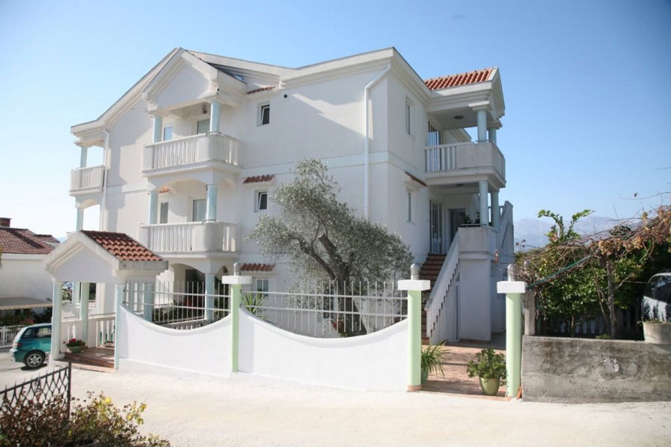 Dreistöckige weiße Villa mit Balkonen und Terrasse in Tivat, Montenegro. Fassade mit Ziegeldach und Olivenbaum.