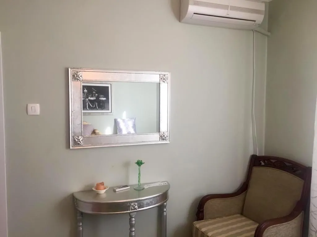 Corner with a silver-framed mirror, console table, and wooden armchair against a light green wall under an AC unit.