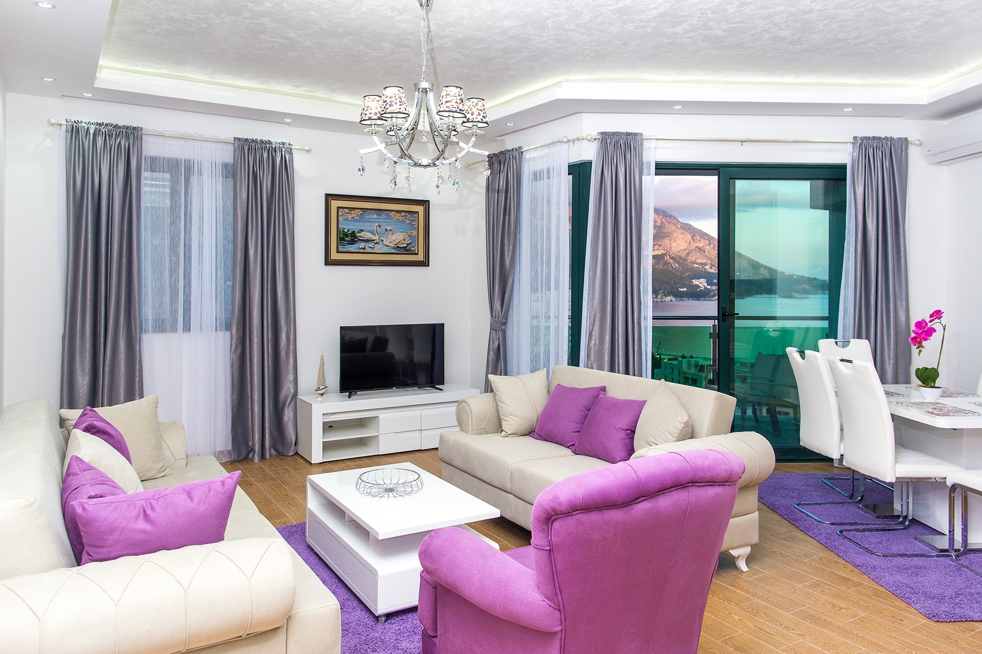 Wohnzimmer in einer Wohnung in Bečići, Montenegro: helle Möbel, violette Akzente und ein Panoramablick auf das Meer und die Berge.