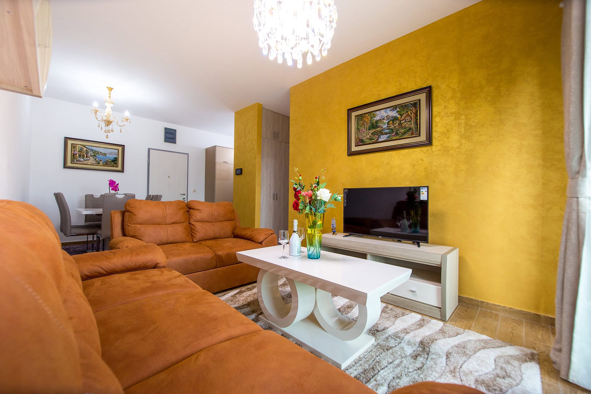 Wohnzimmer in einem Apartment in Bečići, Montenegro: braunes Sofa, gelbe Akzentwand, TV-Bereich und Essgruppe.
