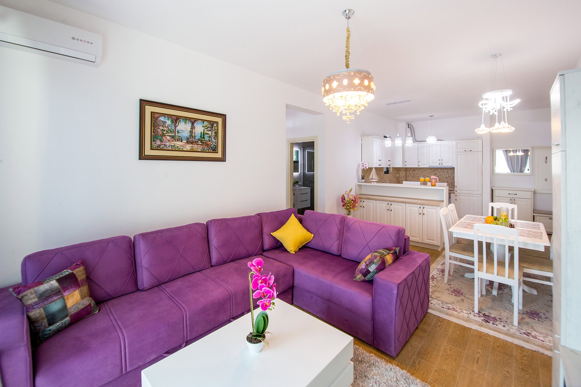 Wohnzimmer mit lilafarbenem Sofa und weißer Küche in einer Wohnung in Bečići, Montenegro. Modernes Interieur mit Essbereich.
