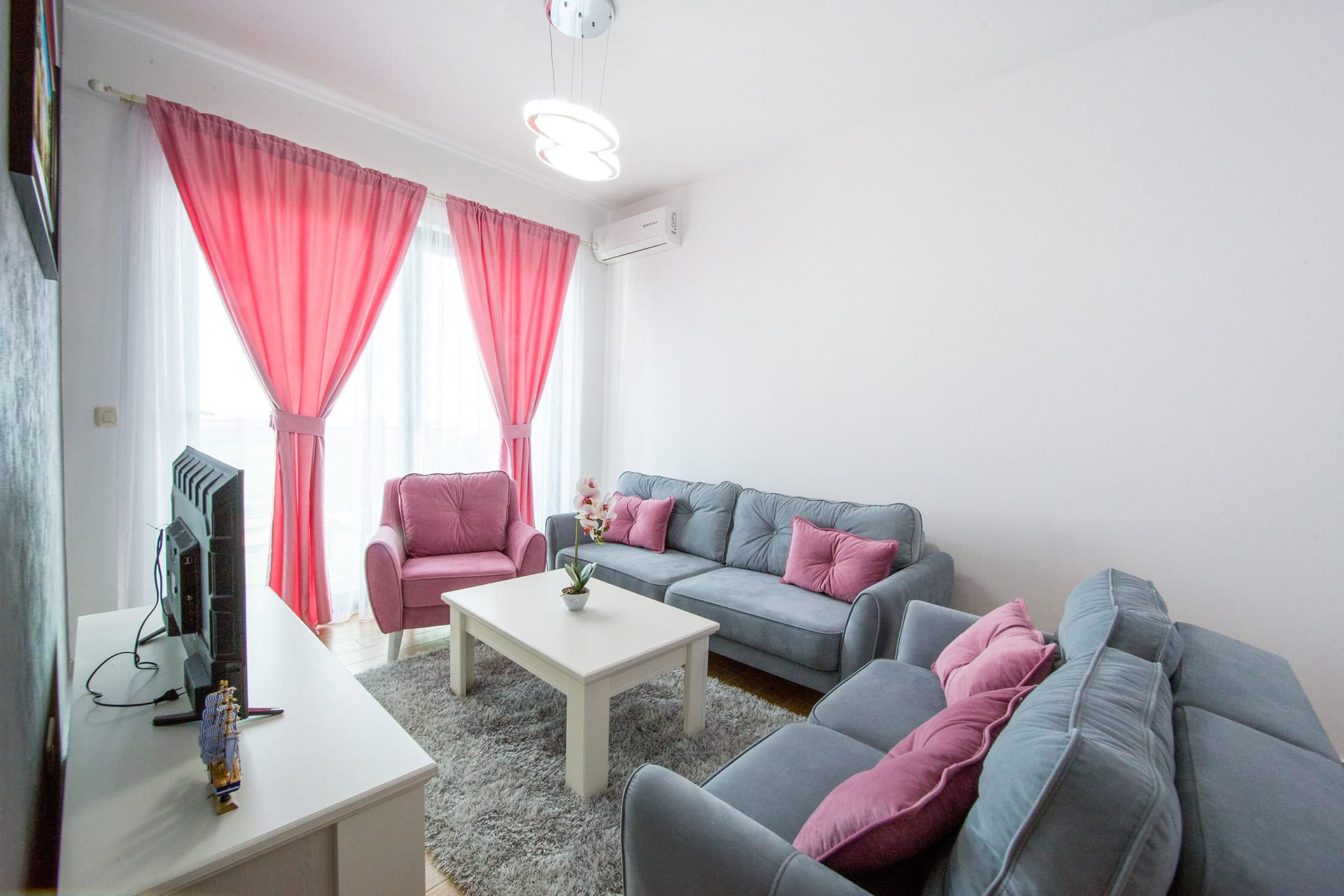 Gemütliches Wohnzimmer in einem Apartment in Bečići, Montenegro: graue Sofas, rosa Sessel, Vorhänge und helle Möbel im Interieur.