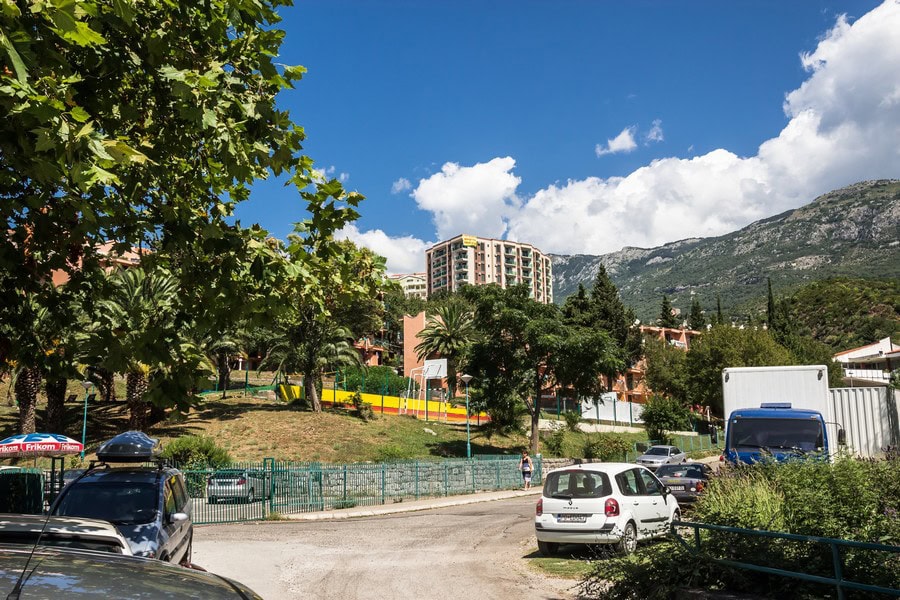 Blick auf die Wohnanlage und die Berge in Bečići, Montenegro: Parkplatz, Grünanlage und mehrstöckige Gebäude vor der Bergkulisse.