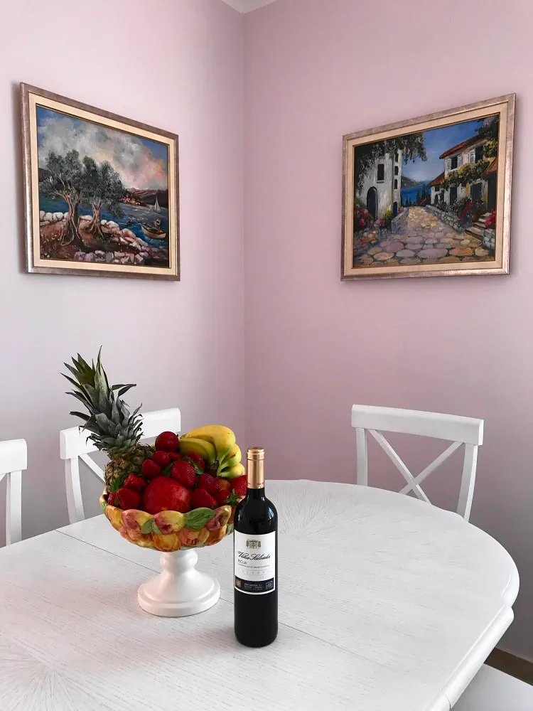 White dining table in the apartment living room in Dobri Vody, Montenegro, with a vase of fruit and wine in front of paintings.