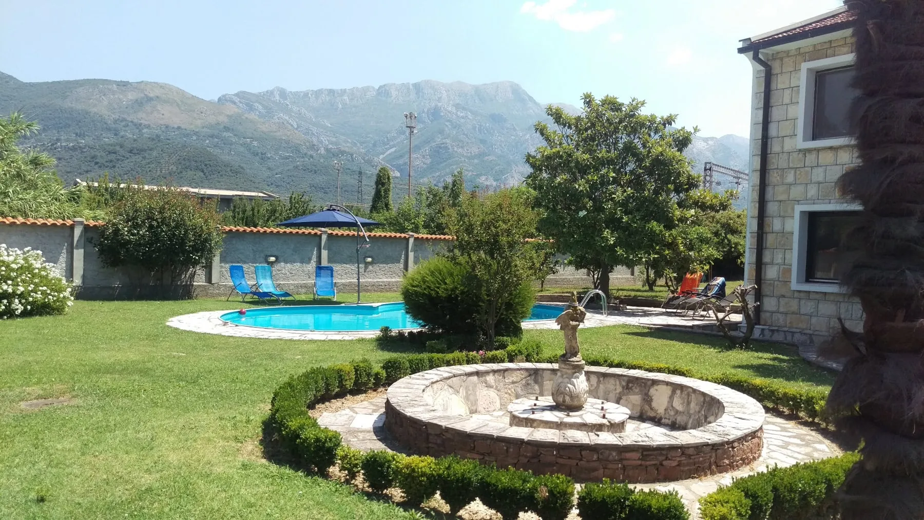 Spacious stone house backyard with pool, fountain, sun loungers, and mountains in the background