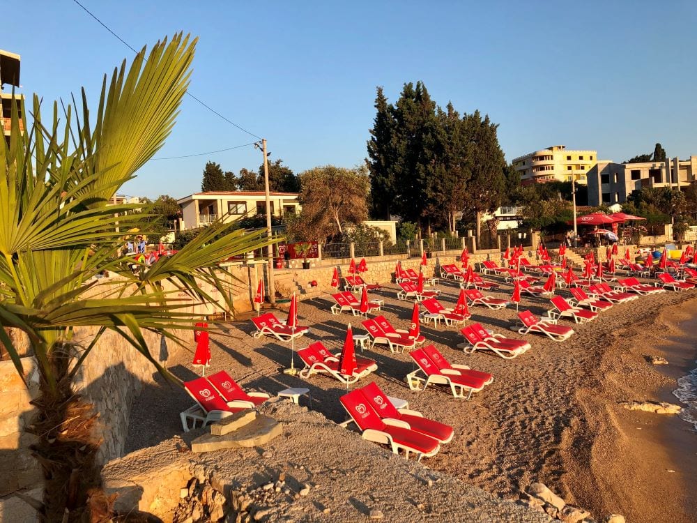 Karadağ, Dobri Vody’de evin yanında kırmızı şezlonglar ve bir palmiye bulunan plaj: deniz kıyısı ve altyapı manzarası.