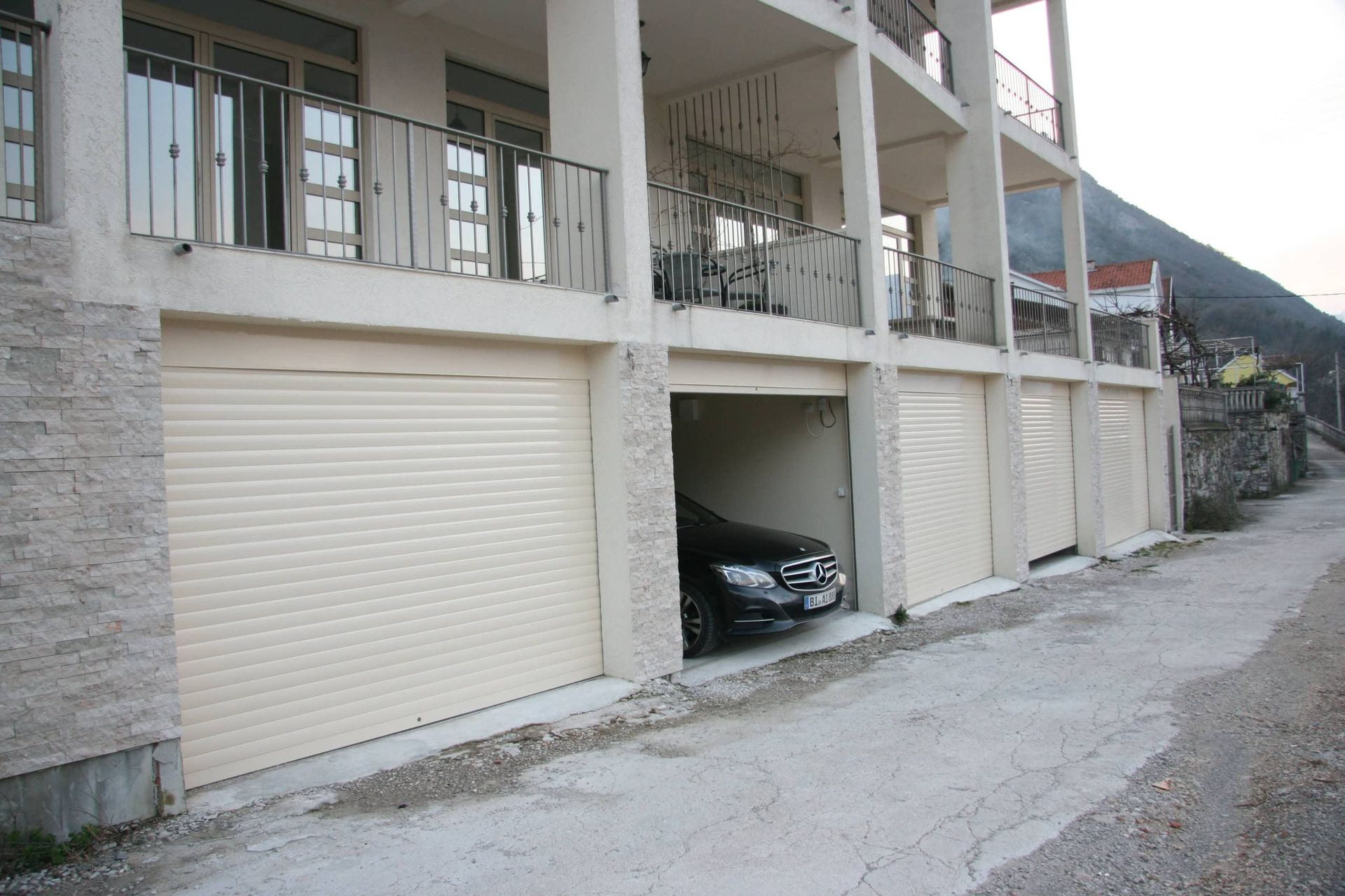 Prčanj, Karadağ’daki evin cephesi: panjurlu garaj bölmeleri, otopark alanı ve ferforje korkuluklu balkonlar.