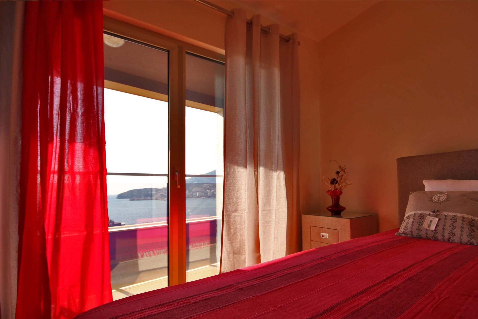 Schlafzimmer in einer Wohnung in Bečići, Montenegro: Meerblick vom Balkon, rote Tagesdecke, Nachttisch und Panoramafenster.