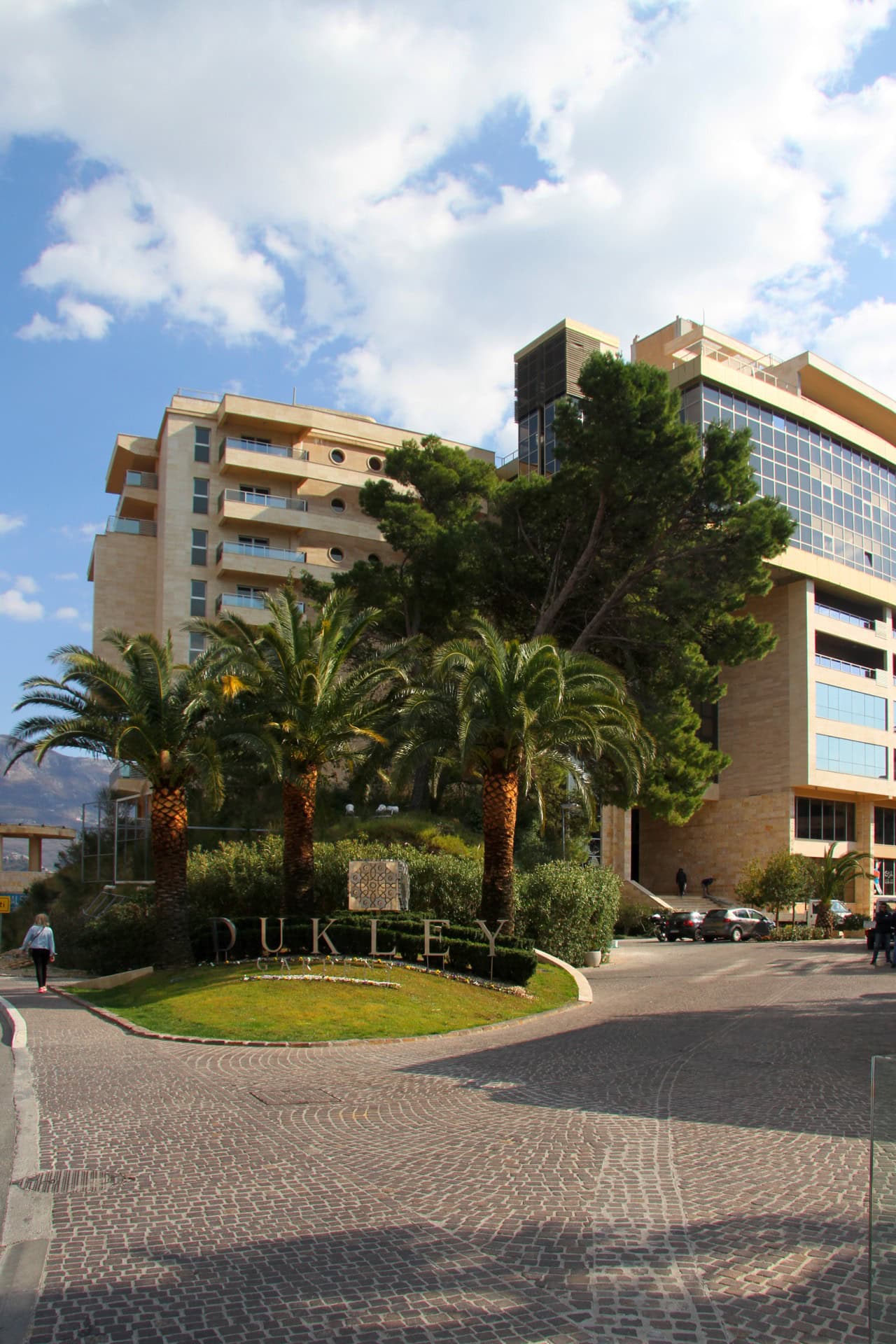 Fassade des Wohnkomplexes Dukley Gardens in Bečići, Montenegro: moderne Architektur, Palmen und gepflegte Außenanlagen.