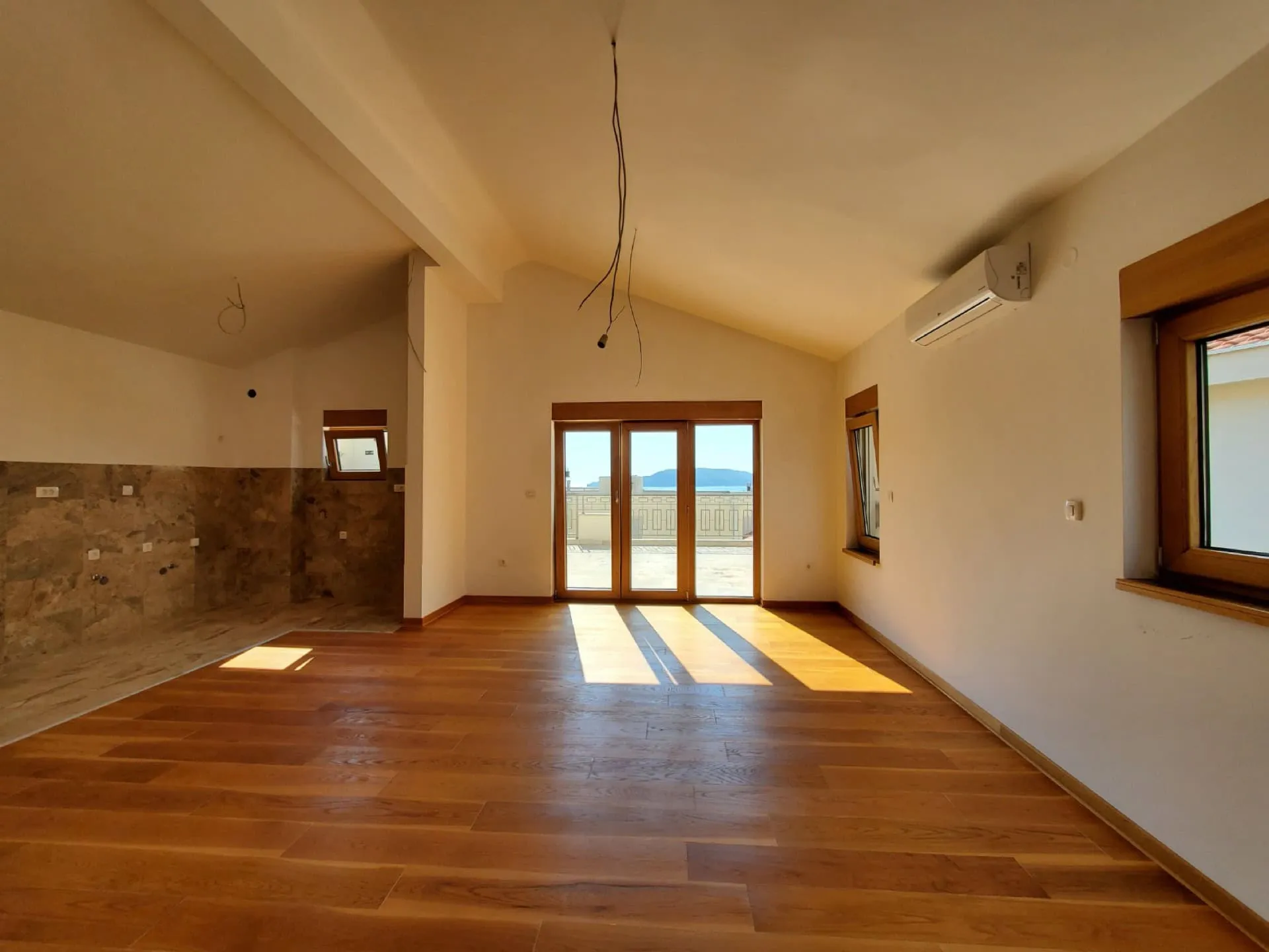 Karadağ, Pržno’daki penthouse’da aydınlık oturma odası: parke zemin, mutfak alanı ve deniz manzaralı terasa çıkış.