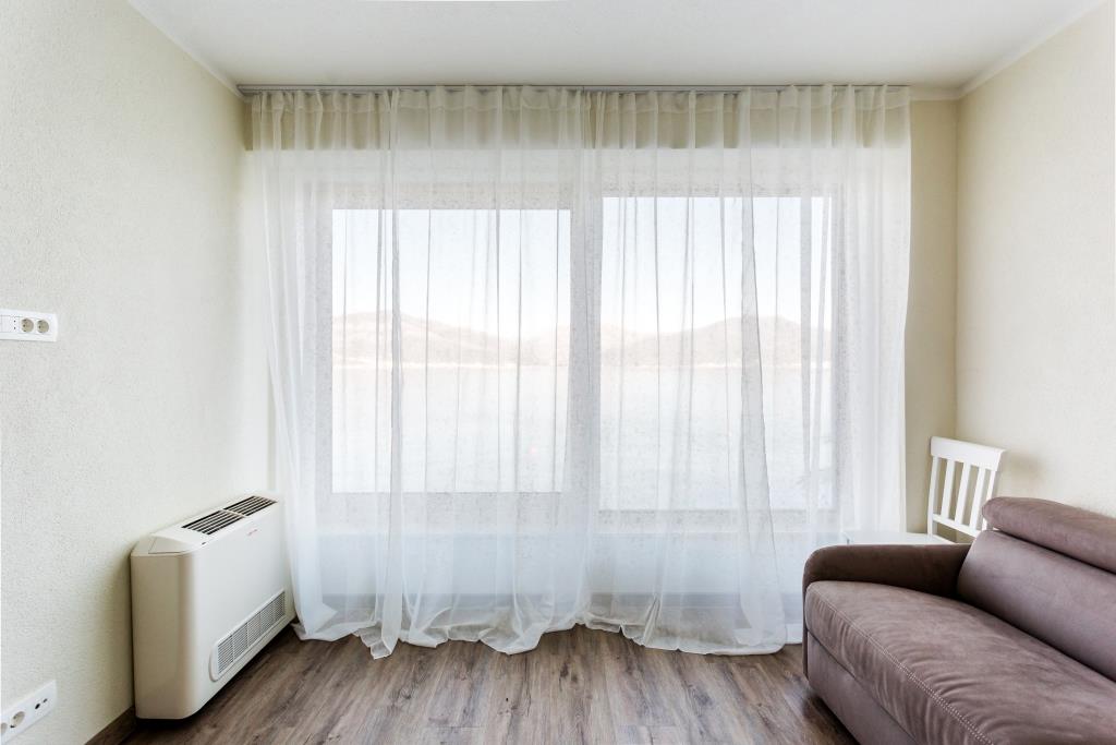 Living room with sea view in a villa, Krašići, Montenegro: brown sofa, light walls, and a panoramic window with sheer curtains.