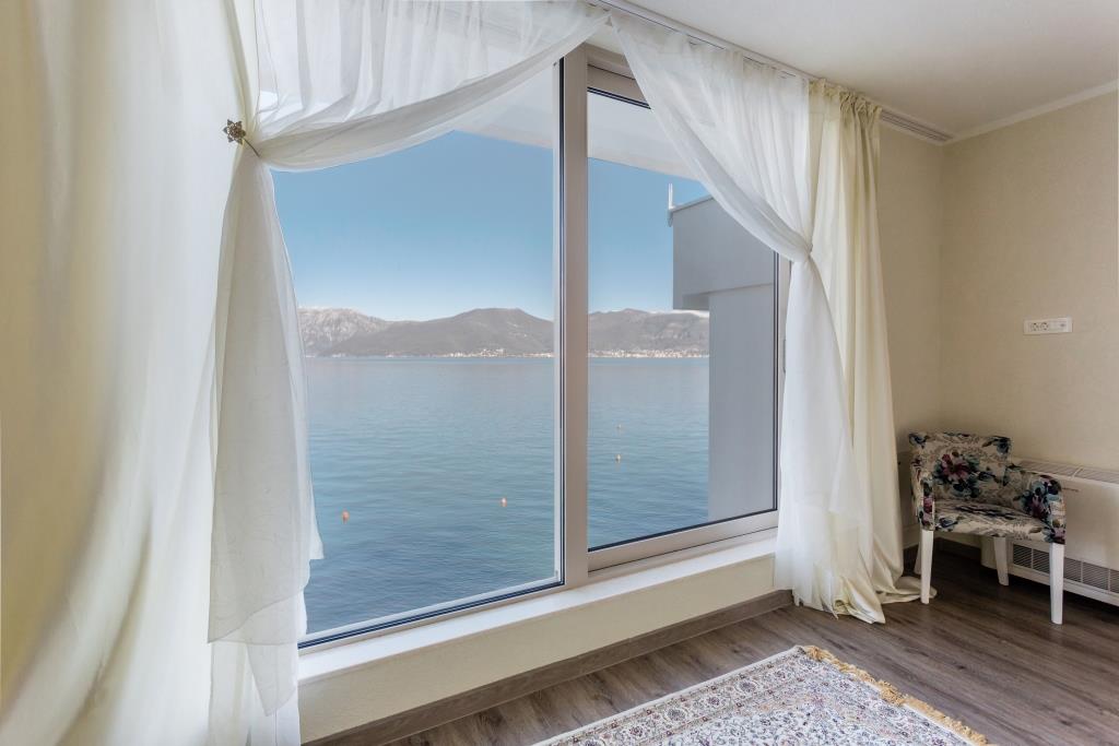 Panoramic window with a view of the sea and mountains in the villa living room in Krašići, Montenegro, with a cozy armchair and light curtains.