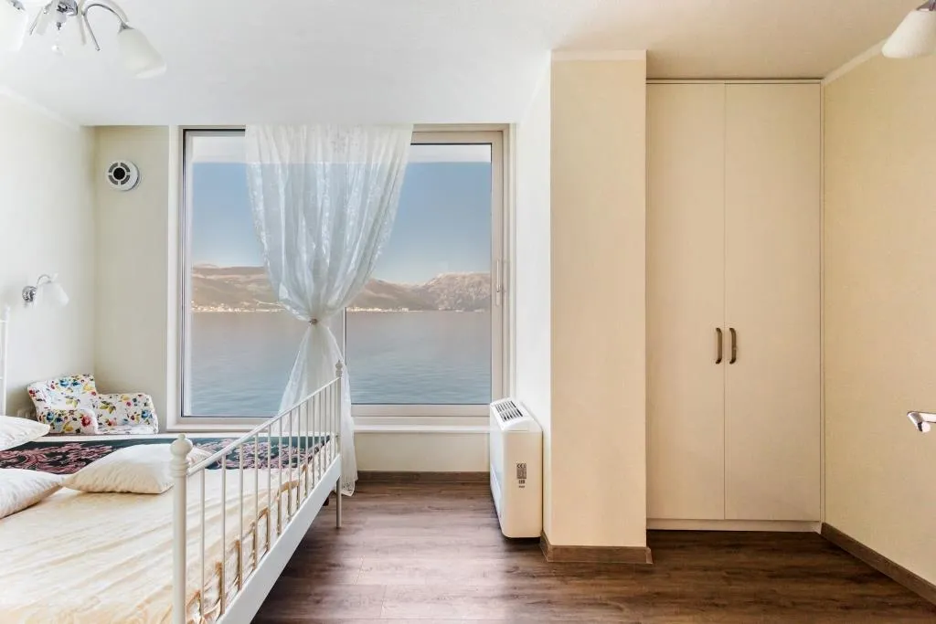 Bedroom with sea view in a villa, Krašići, Montenegro: white bed, built-in wardrobe, and panoramic window overlooking the bay.