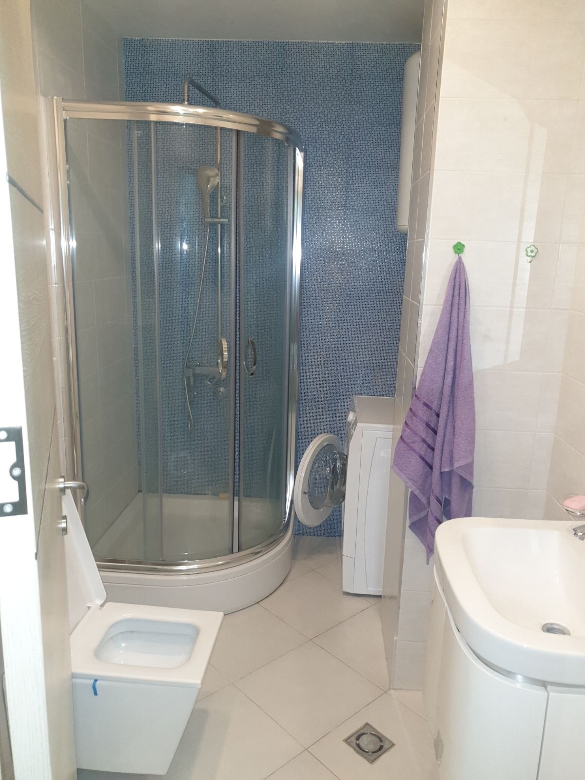 Badezimmer in einer Wohnung in Budva, Montenegro: Duschkabine, Waschmaschine, Waschbecken und helle Fliesen.