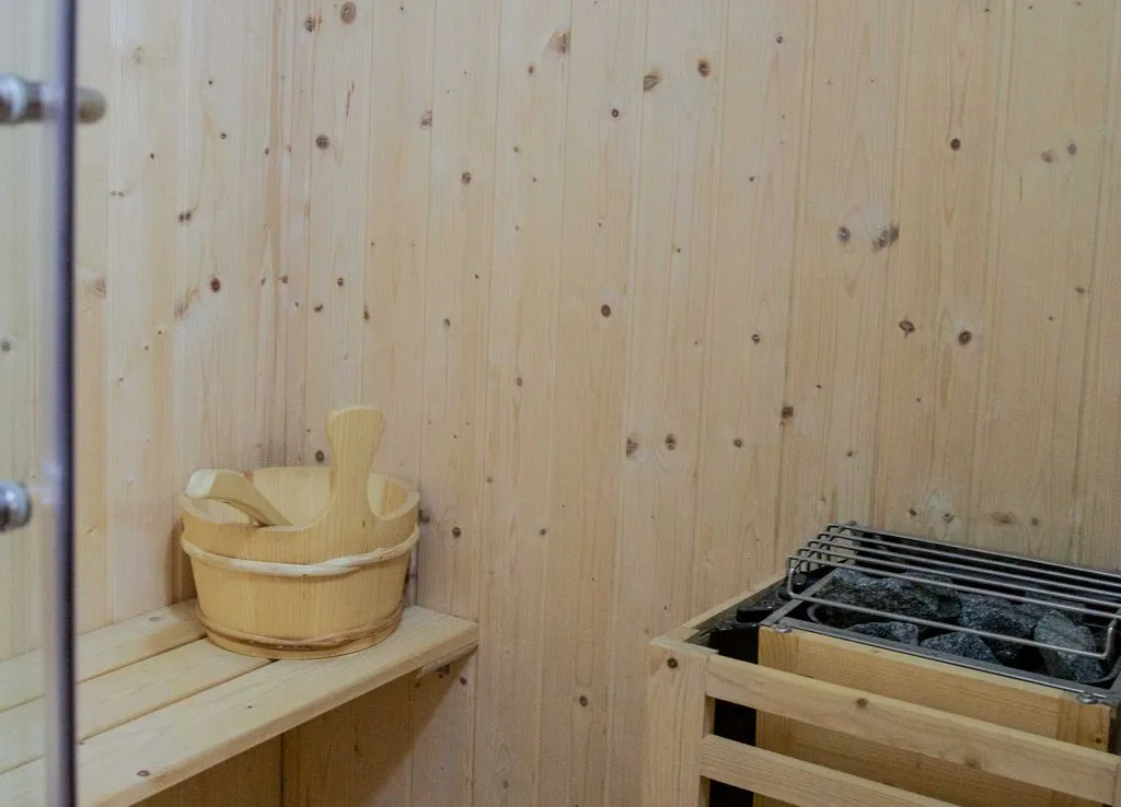Interijer saune: drvene stene, bench, drvena kanta sa lopatom i peć sa kamenjem.
