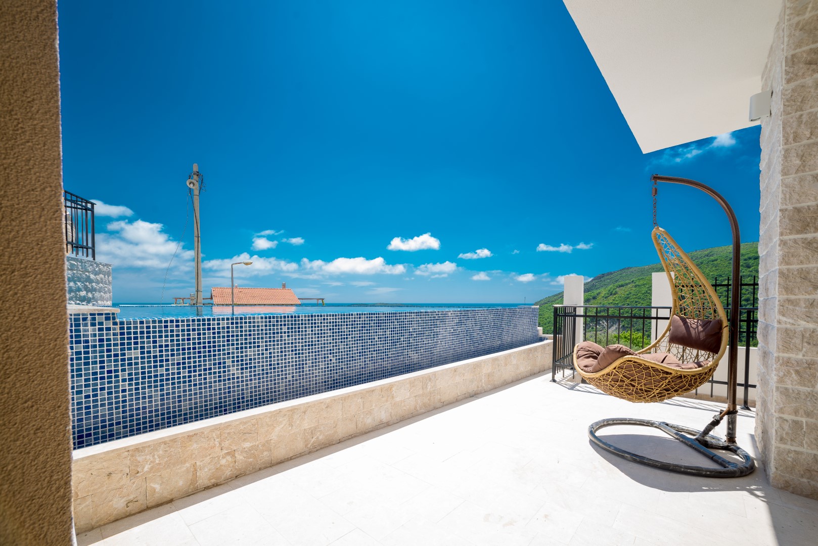 Terrasse der Villa in Bečići, Montenegro: Infinity-Pool mit blauer Mosaikverkleidung, geflochtener Kokonstuhl und Meerblick.