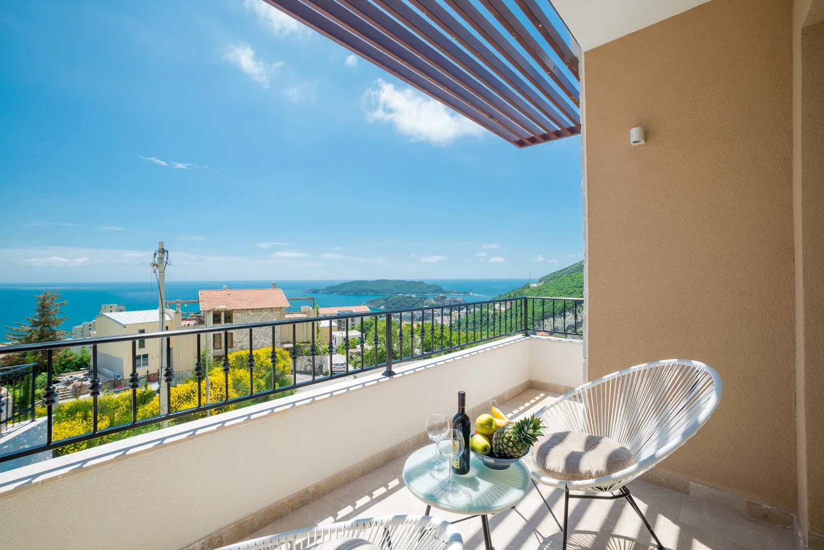 Sonniger Balkon mit weißen Korbstühlen, einem kleinen Glastisch mit Wein und Obst sowie Blick auf das Meer und die Stadt.
