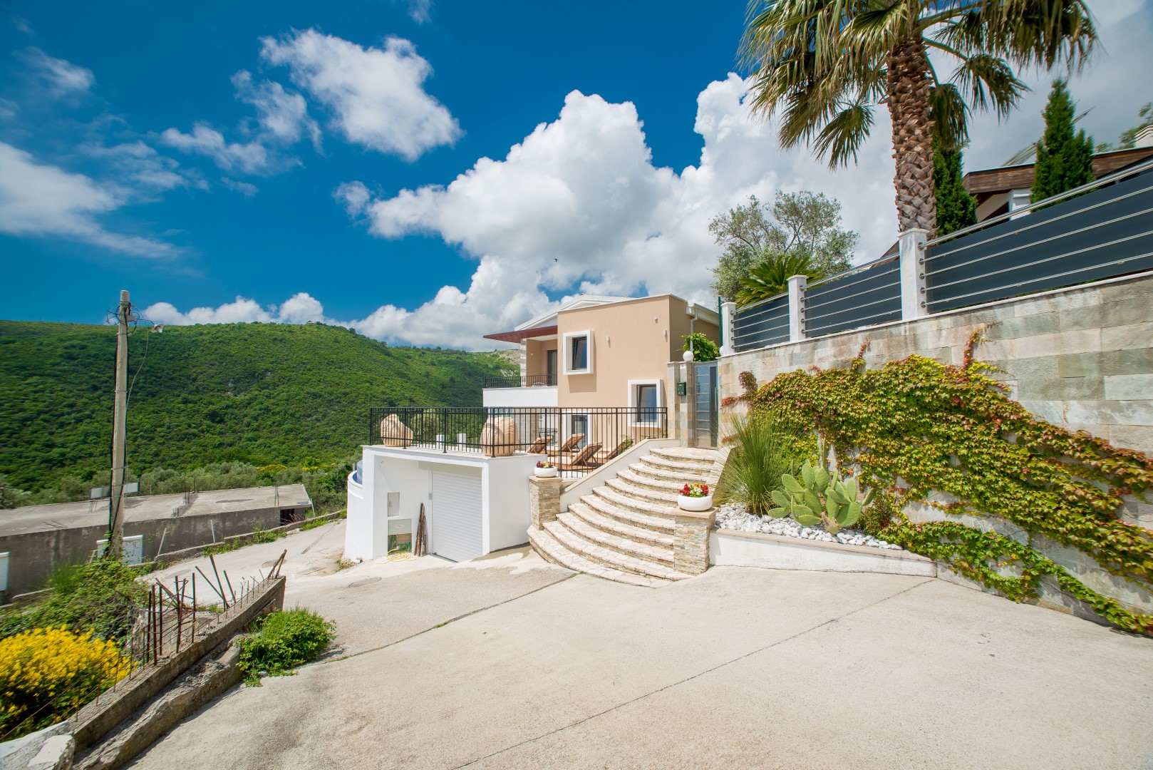 Moderne Villa in Bečići, Montenegro: Fassade mit Terrasse, Steintreppe, Palme und Blick auf grüne Berge.