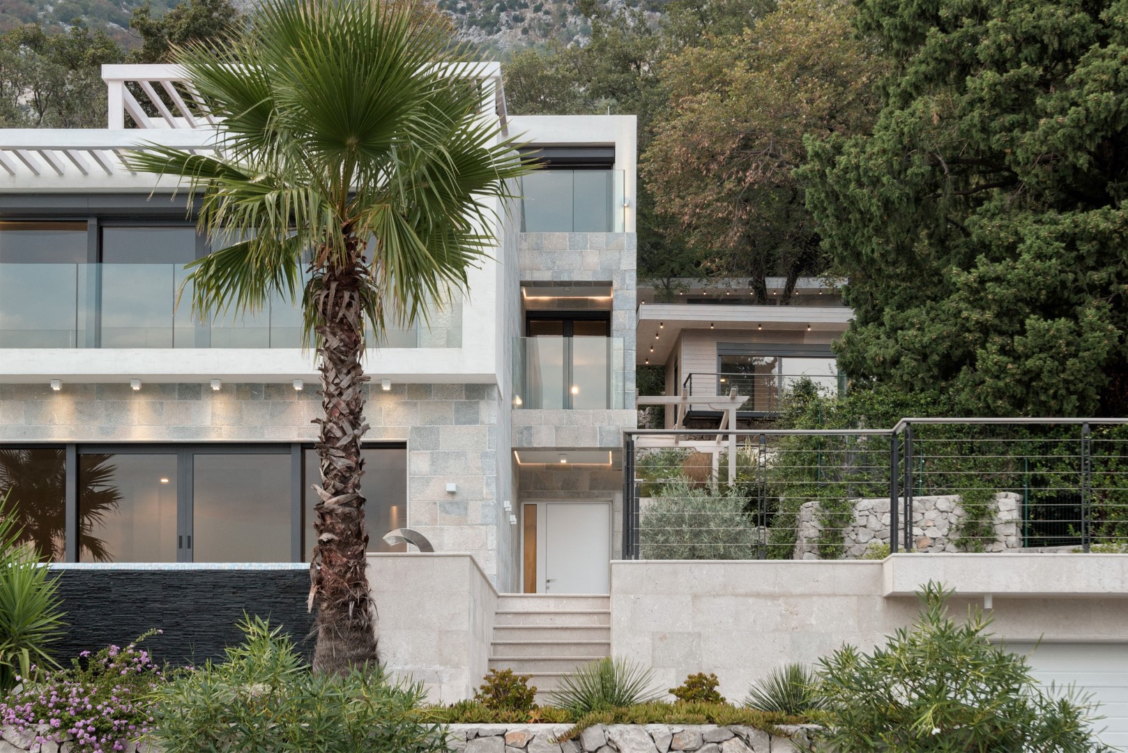 Karadağ, Strp’te modern villa: taş cepheli, panoramik pencereli, teraslı ve dağ manzarası fonunda palmiye ağacı.