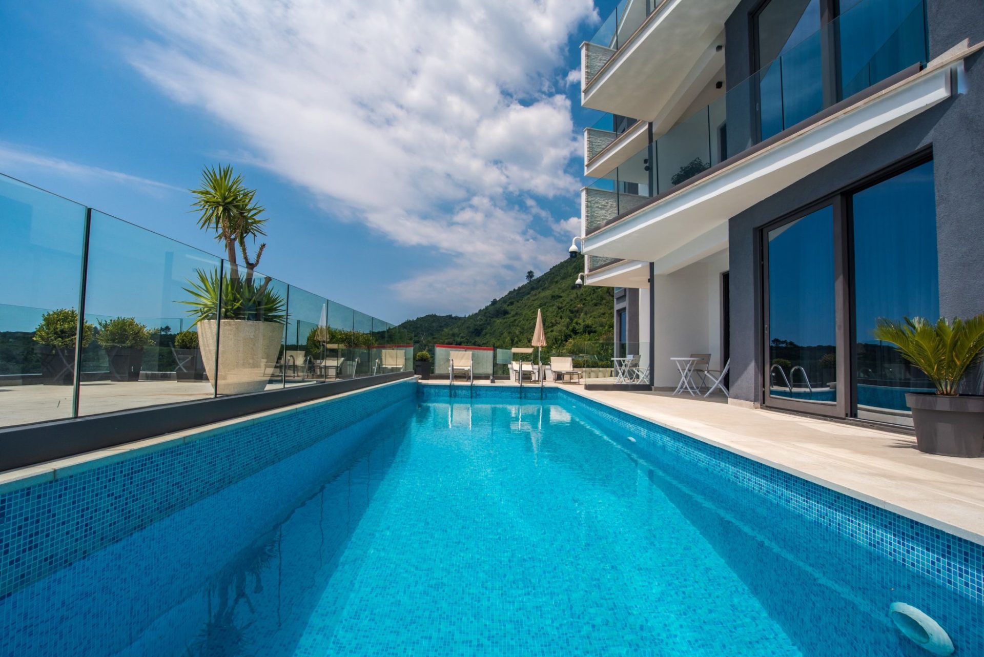 Offener Pool mit blauen Fliesen auf der Terrasse einer modernen Villa in Budva, Montenegro, mit Blick auf die Berge und Glasgeländer.