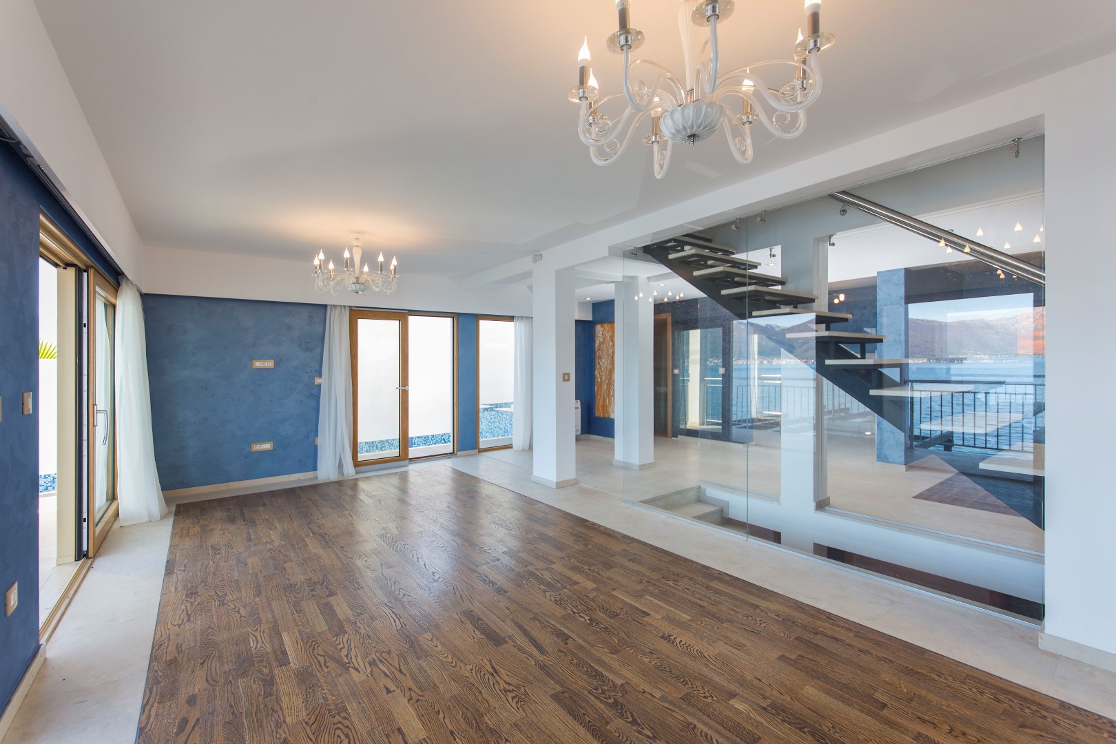 Spacious villa living room in Krašići, Montenegro: wooden floor, glass staircase, and panoramic sea view.