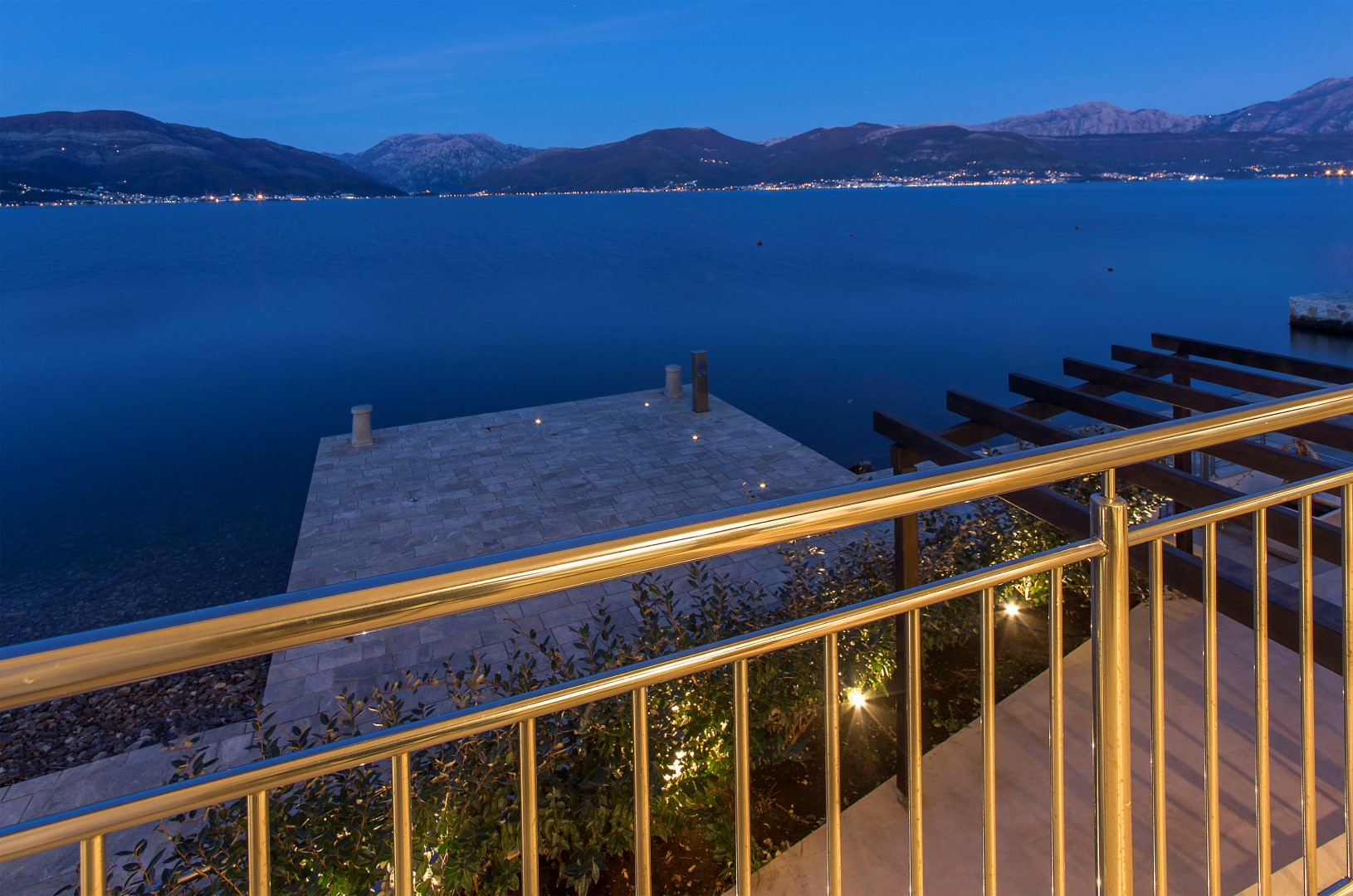 View from the villa balcony in Krašići, Montenegro: a private stone pier, the sea, and the night lights of the mountain coast.