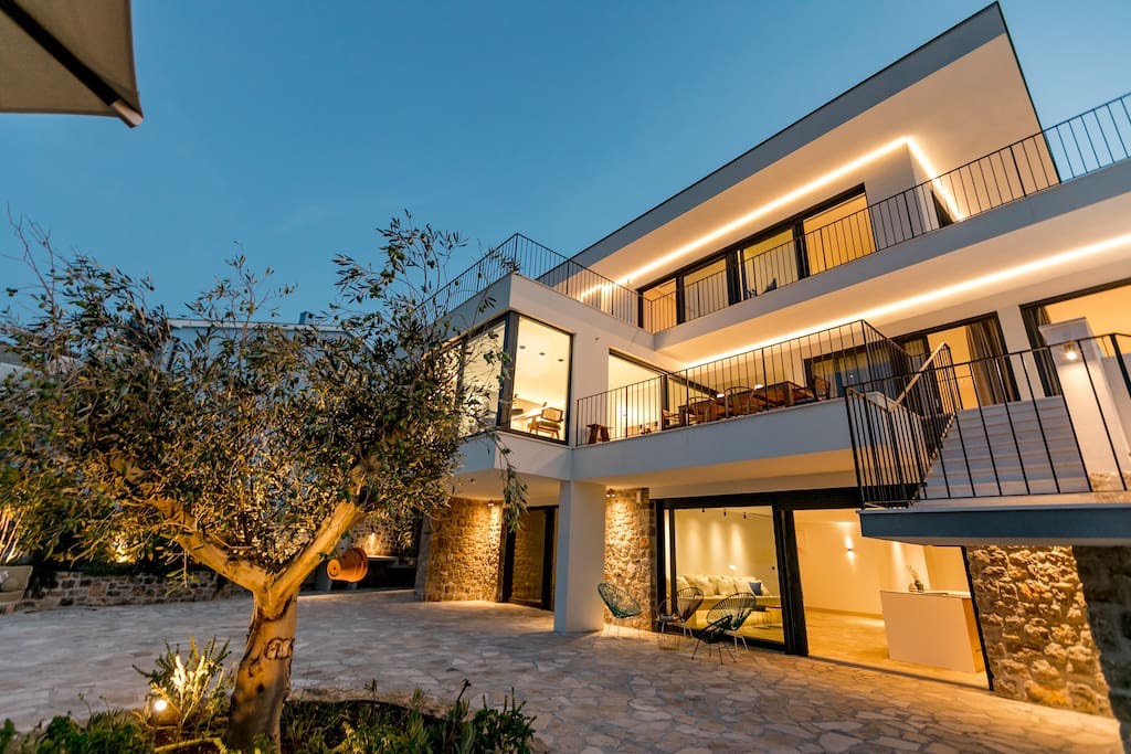 Moderne Villa in Luštica, Montenegro: Fassade mit Beleuchtung, Steinverkleidung, Terrasse und Olivenbaum im Hof.