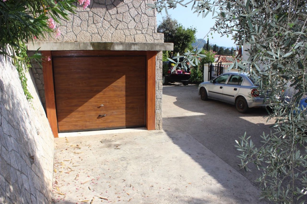 Garage mit braunem Tor und Parkplatz an einer Villa in Budva, Montenegro: Steinfassade und Olivenbaum.