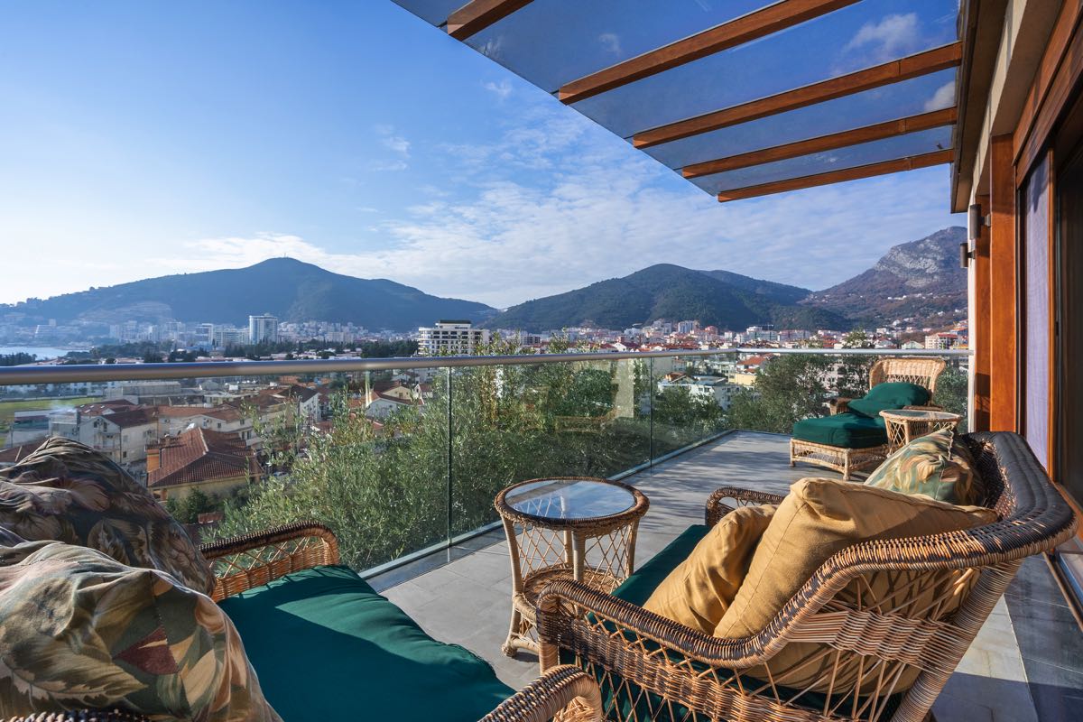 Terrasse einer Villa in Budva, Montenegro: geflochtene Möbel, Glasgeländer und Panoramablick auf die Stadt und die Berge.