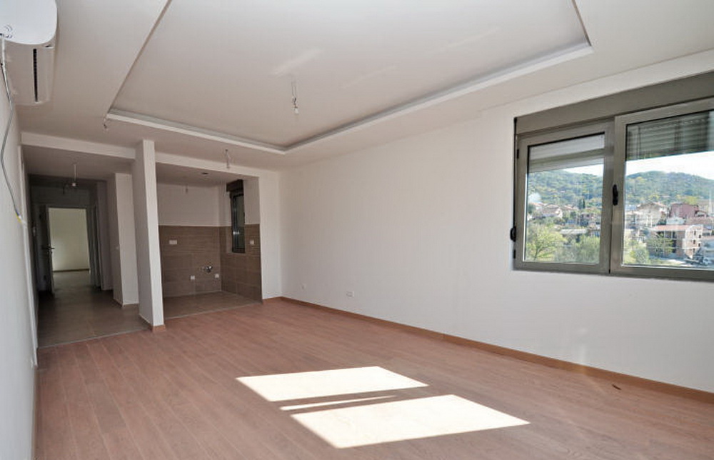Helles Wohnzimmer mit Laminat und Küchenbereich in einer neuen Wohnanlage in Tivat, Montenegro, mit Bergblick aus dem Fenster.