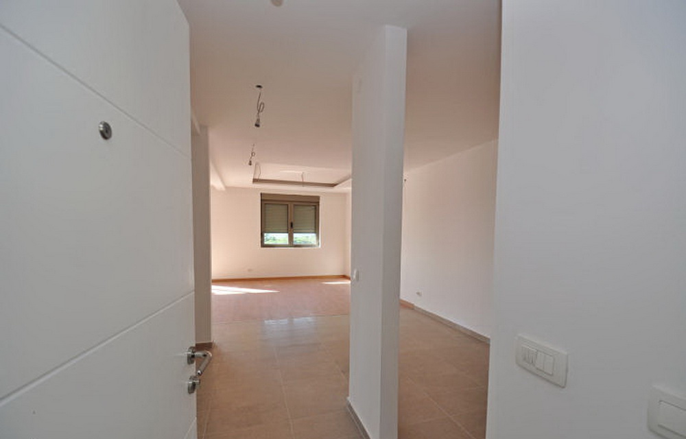 Helles Wohnzimmer in einem Neubau in Tivat, Montenegro: weiße Wände, Fliesenboden und ein Fenster mit Jalousien in einer leeren Wohnung.