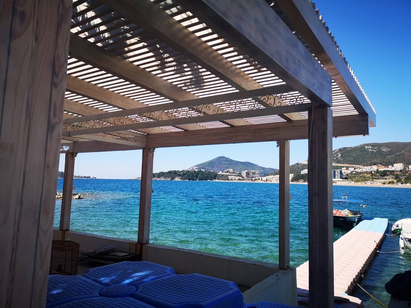 Terrasse d’un local commercial avec vue sur la mer et le quai à Rafailovici, Monténégro, sous un auvent en bois.