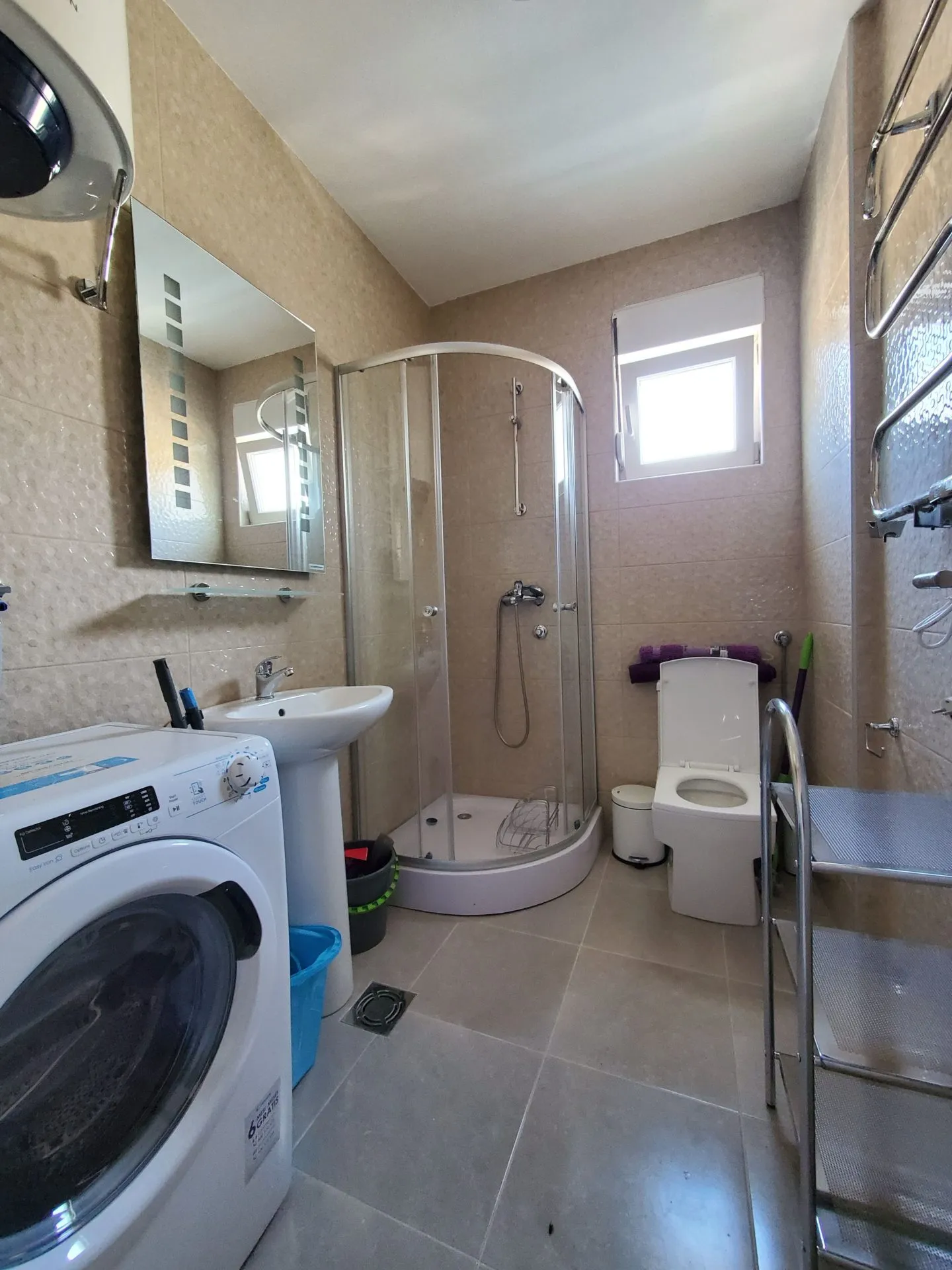 Badezimmer in einer Wohnung in Budva, Montenegro: Duschkabine, Waschmaschine, Waschbecken und helle Fliesen.