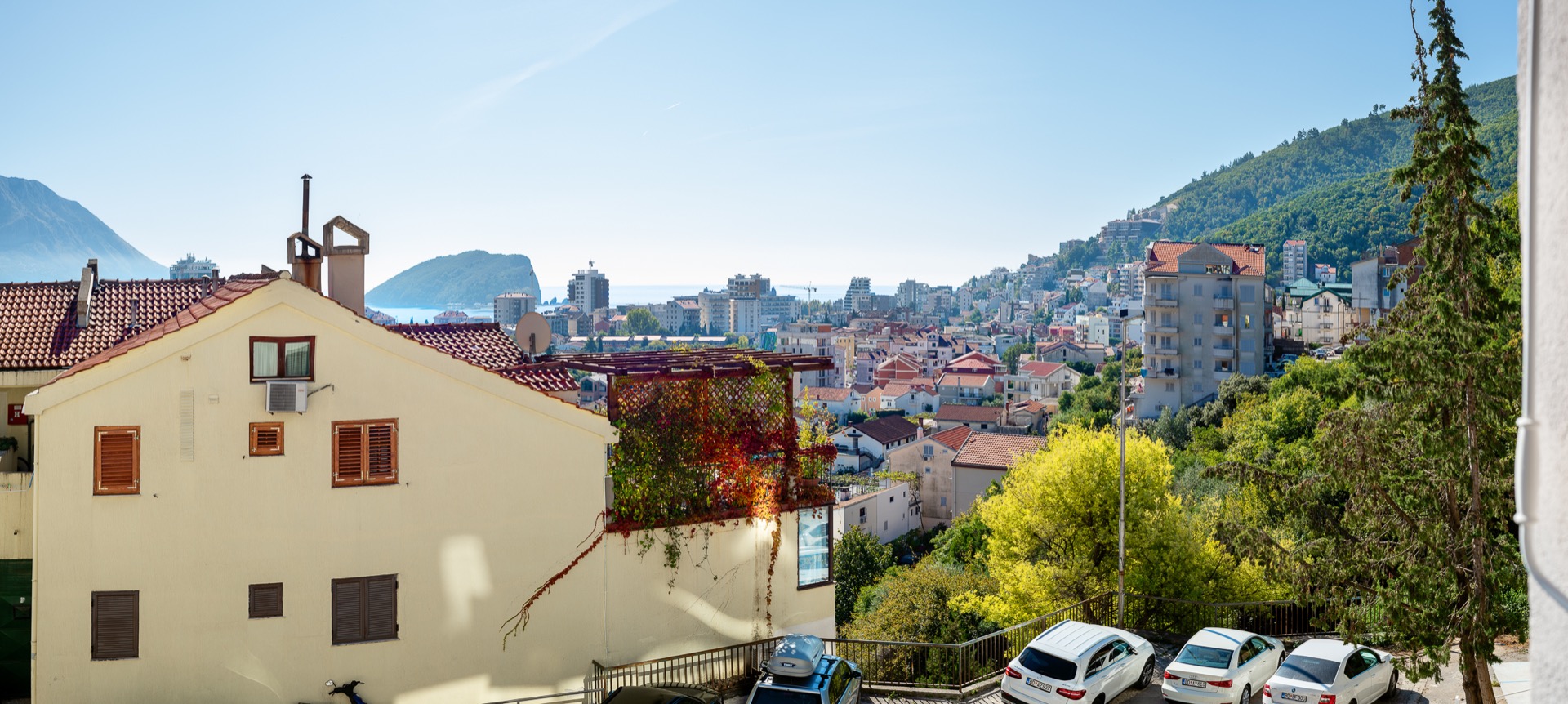 Panoramablick auf die Stadt Budva, Montenegro, mit Terrasse, grünen Bäumen und der Küste am Horizont.