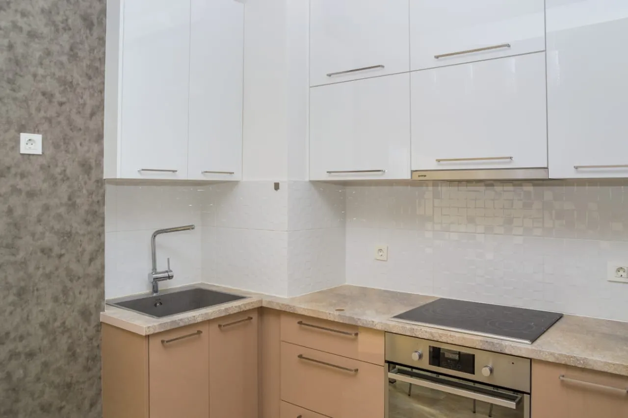Beyaz üst ve bej alt dolaplar, lavabo, ocak ve gömme fırın olan modern bir mutfak köşesi.