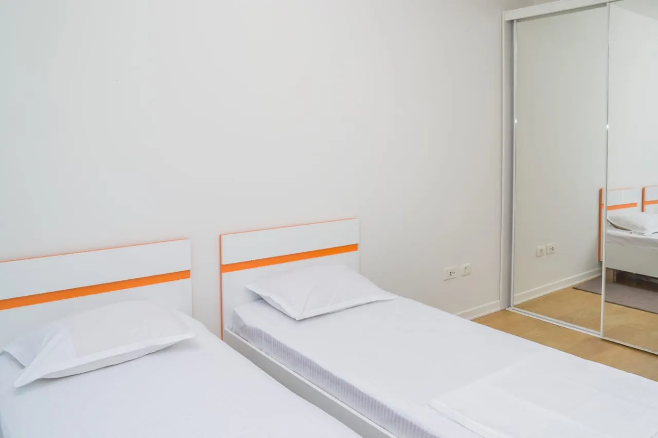 Başlıklarda turuncu detayları olan beyaz çamaşırlı iki tek kişilik yatak ve yanındaki mirror dolap.
