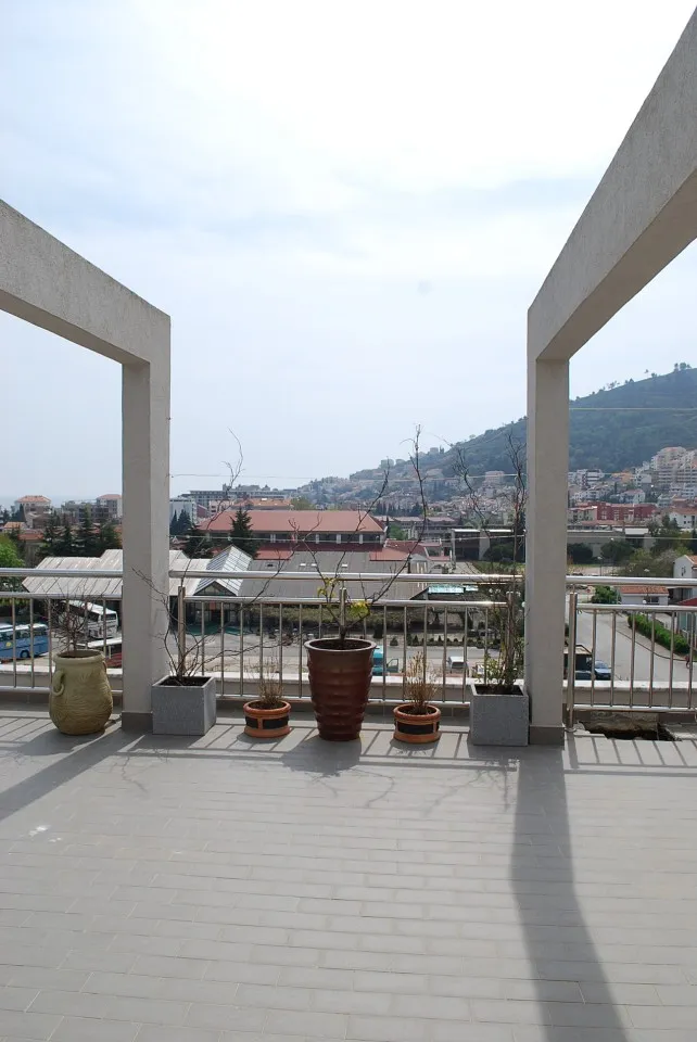Open balcony with gray tiles, metal railing, and decorative potted plants overlooking a cityscape and distant hills under a bright sky.