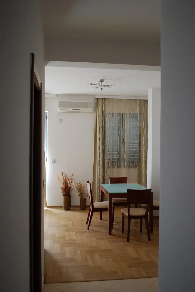 View from a hallway into a room with parquet flooring, a dining table, chairs, and a window with curtains.