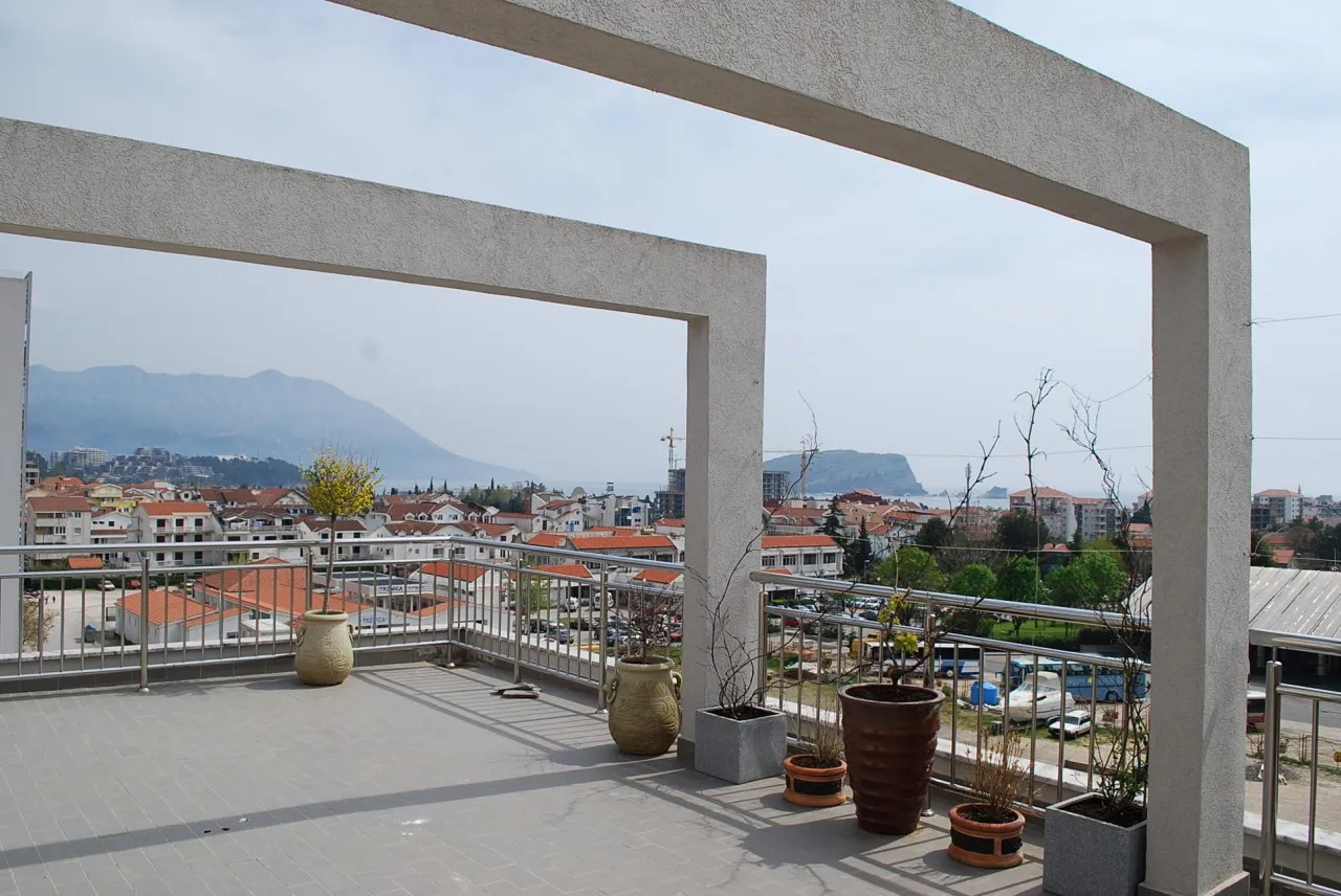 Open balcony with concrete columns and metal railing overlooking a coastal town and mountains.